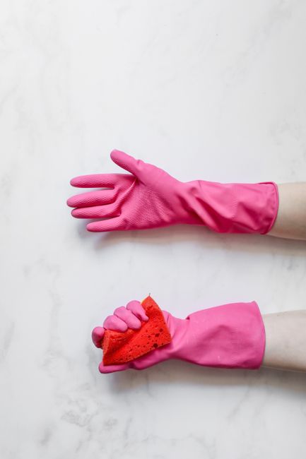 A person wearing orange gloves and white protective clothing is using a modern, upright vacuum cleaner to clean a smooth, grey tiled floor in a contemporary living space. The vacuum cleaner features a