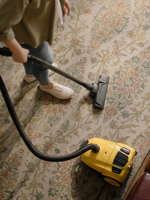 A person operating a yellow portable carpet cleaning machine on a patterned area rug in a room with natural daylight. The machine has a black handle and control panel, with visible vent grilles on the