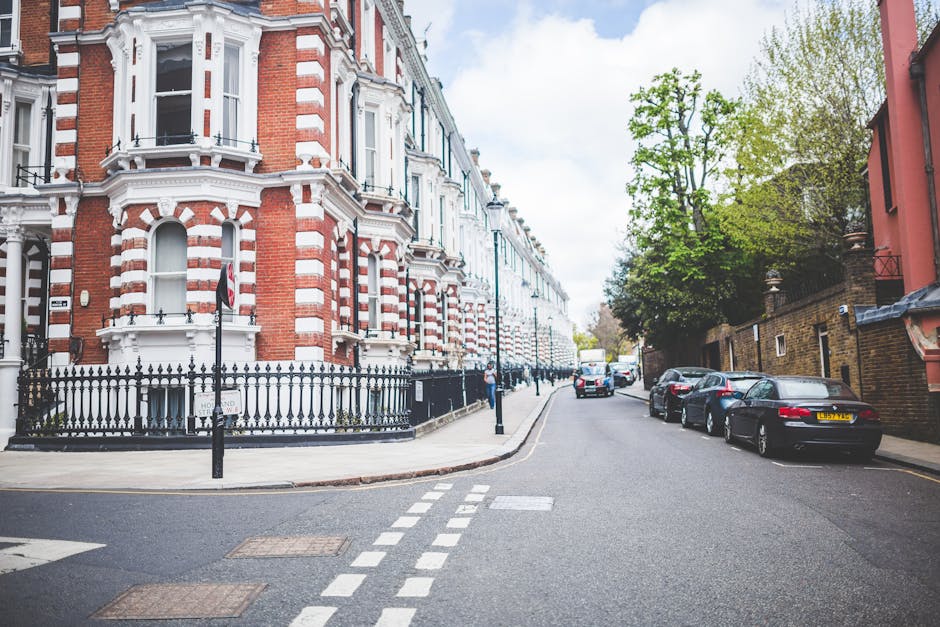 A street scene on Upper Richmond Road in Putney featuring Victorian-style residential buildings with red-brick facades and white decorative trim. The buildings have large windows and ornate architectural details, with a black wrought-iron fence bordering the sidewalk. Several parked cars line the street on the right, including a black coupe and a silver sedan. The street is paved with asphalt and has white dashed and solid lane markings. Trees with fresh green leaves are visible on the right side, providing partial shade. The scene is illuminated by natural daylight, emphasizing the clean, well-maintained appearance of both the buildings and the street, representative of the area where Cleaners Putney offers domestic cleaning services such as surface cleaning and deep cleaning for residential properties in Putney.