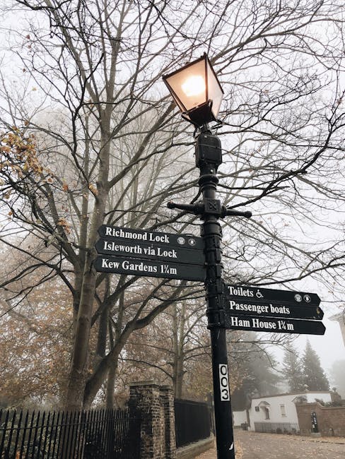 A black metal street lamp post with a lit lantern at the top, situated amidst leafless trees on a foggy day. Attached to the post are multiple black directional signs indicating nearby locations, including 'Richmond Lock,' 'Isleworth via Lock,' 'Kew Gardens 1/2 m,' 'Toilets & Passenger boats,' and 'Ham House 1/4 m.' The background features a black metal fence, a small white building, and distant fog-covered trees, creating an atmospheric scene. This image exemplifies outdoor street scene elements with signage and natural surroundings, suitable for illustrating location guidance or urban environment imagery, consistent with the context of residential and commercial cleaning services provided by Cleaners Putney on their website page titled 'Upper Richmond Road End-of-Tenancy Cleaners in Putney, PUTNEY'.
